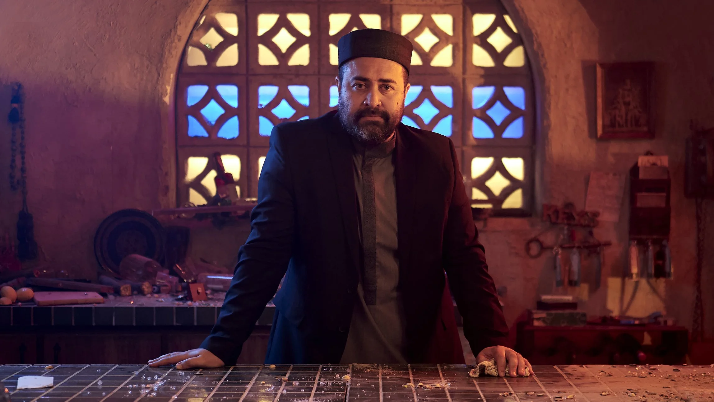 A man with a beard wearing a black hat and dark clothing stands in a dimly lit room with colorful lighting. He is leaning on a tiled counter with objects and debris scattered around. The background features decorative wall openings with blue and yell