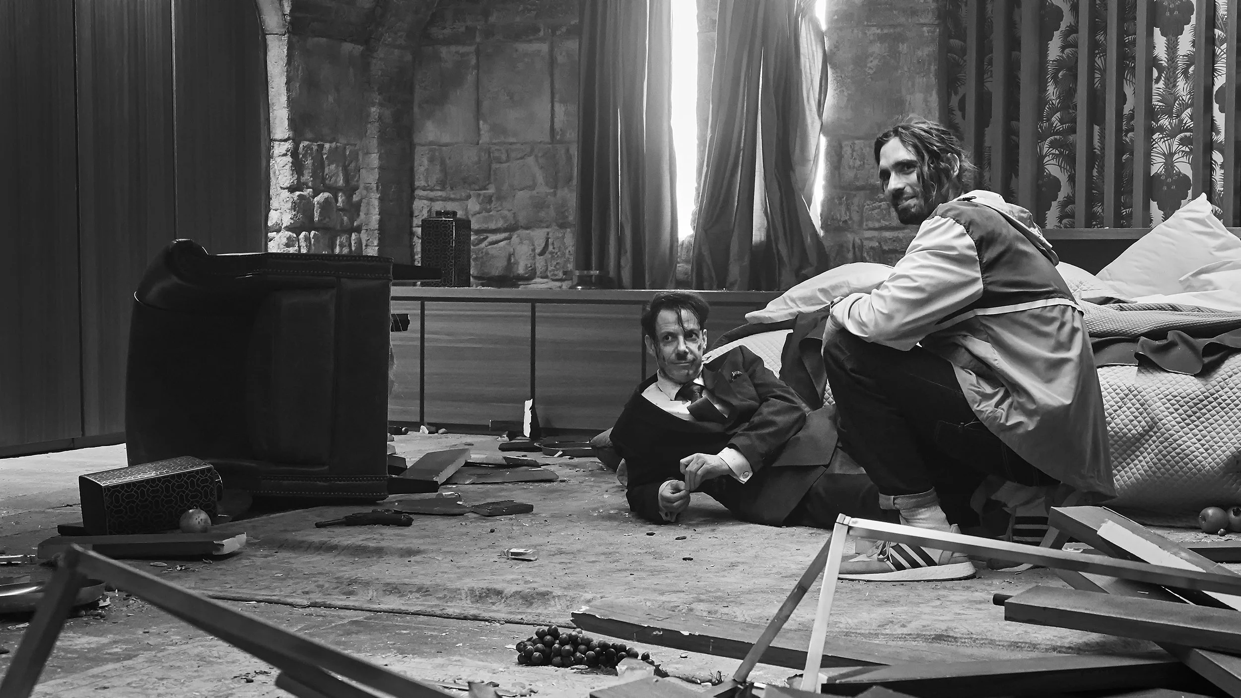Two men are inside a room with stone walls and a window with curtains. One man is sitting on the floor, looking at the camera, wearing a suit and tie. The other man is crouched next to him, smiling, wearing a jacket. The room is cluttered with variou