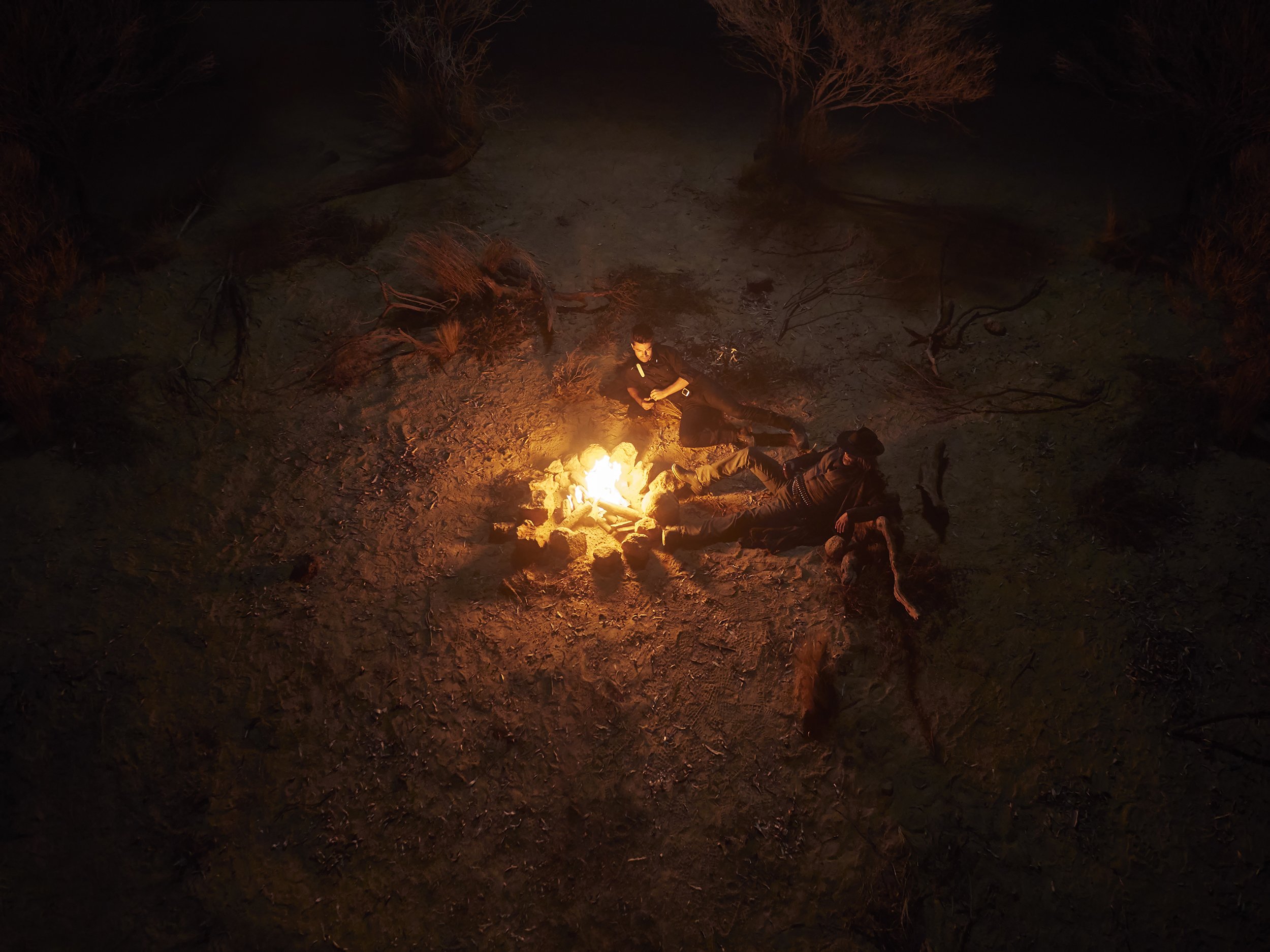 Two people sitting near a campfire at night in a desert area with dry trees and open ground.