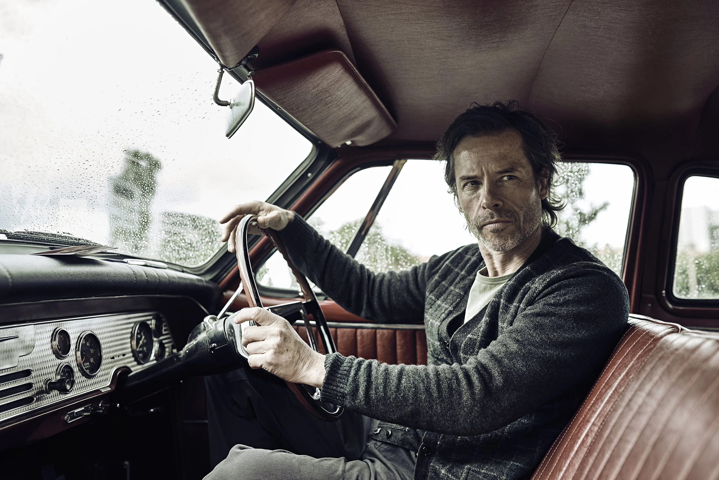 sitting in the driver's seat of a vintage car, holding the steering wheel, with a serious expression on his face. The car interior features brown leather seats and a dashboard with multiple gauges. The rainy weather outside causes raindrops on the wi
