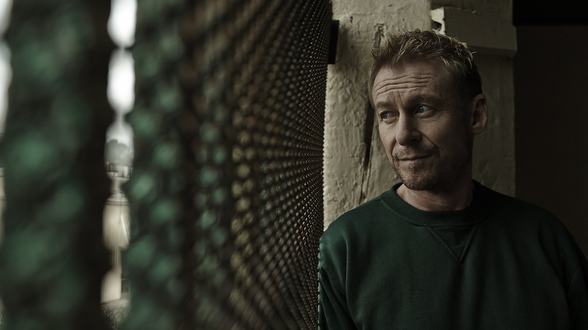 A man with short, curly hair and a beard looking to his left, leaning against a stone wall in a dimly lit space, with a black textured surface nearby.