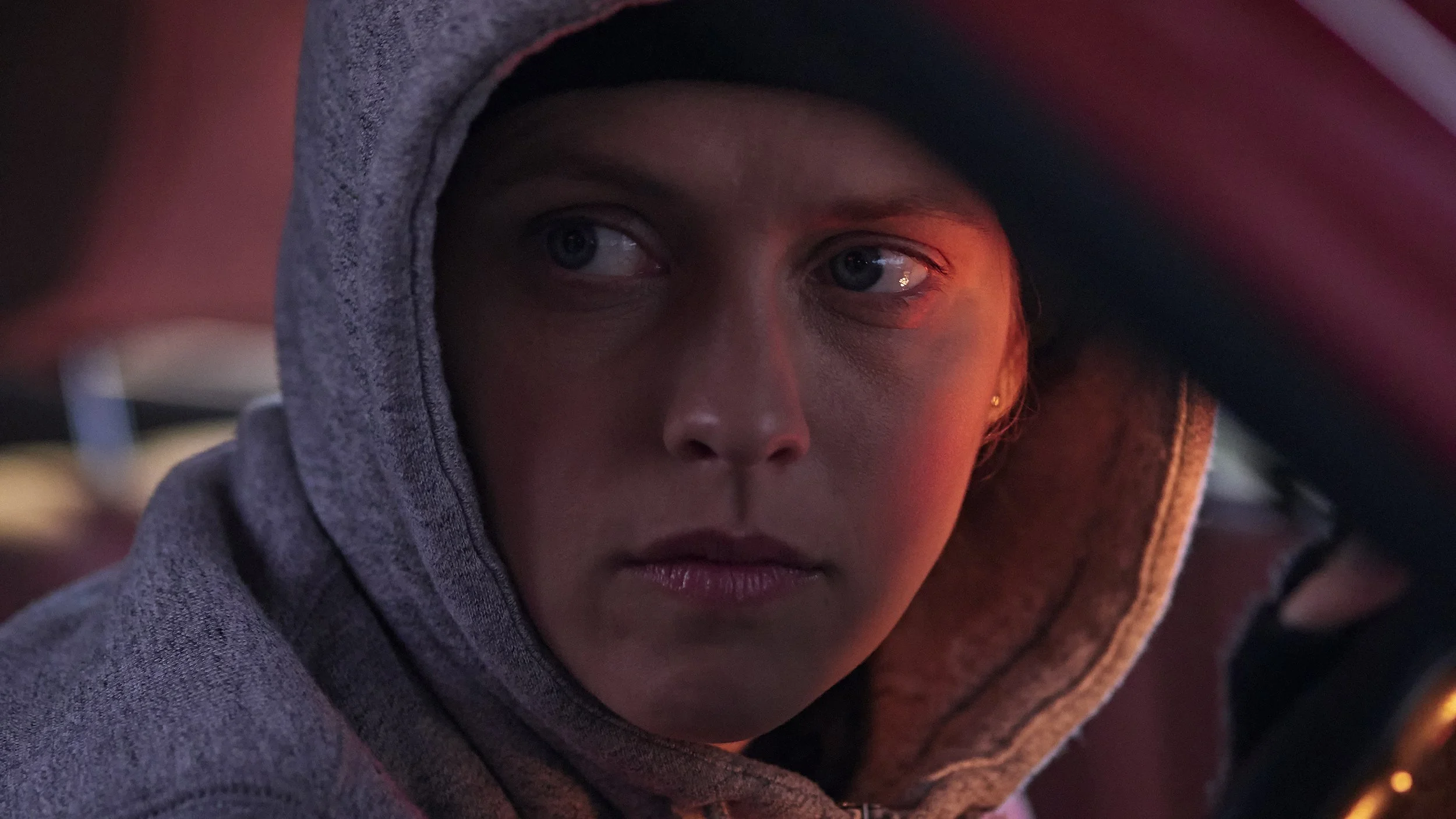 Close-up of a young person wearing a gray hoodie, with a serious expression, illuminated by red lighting.