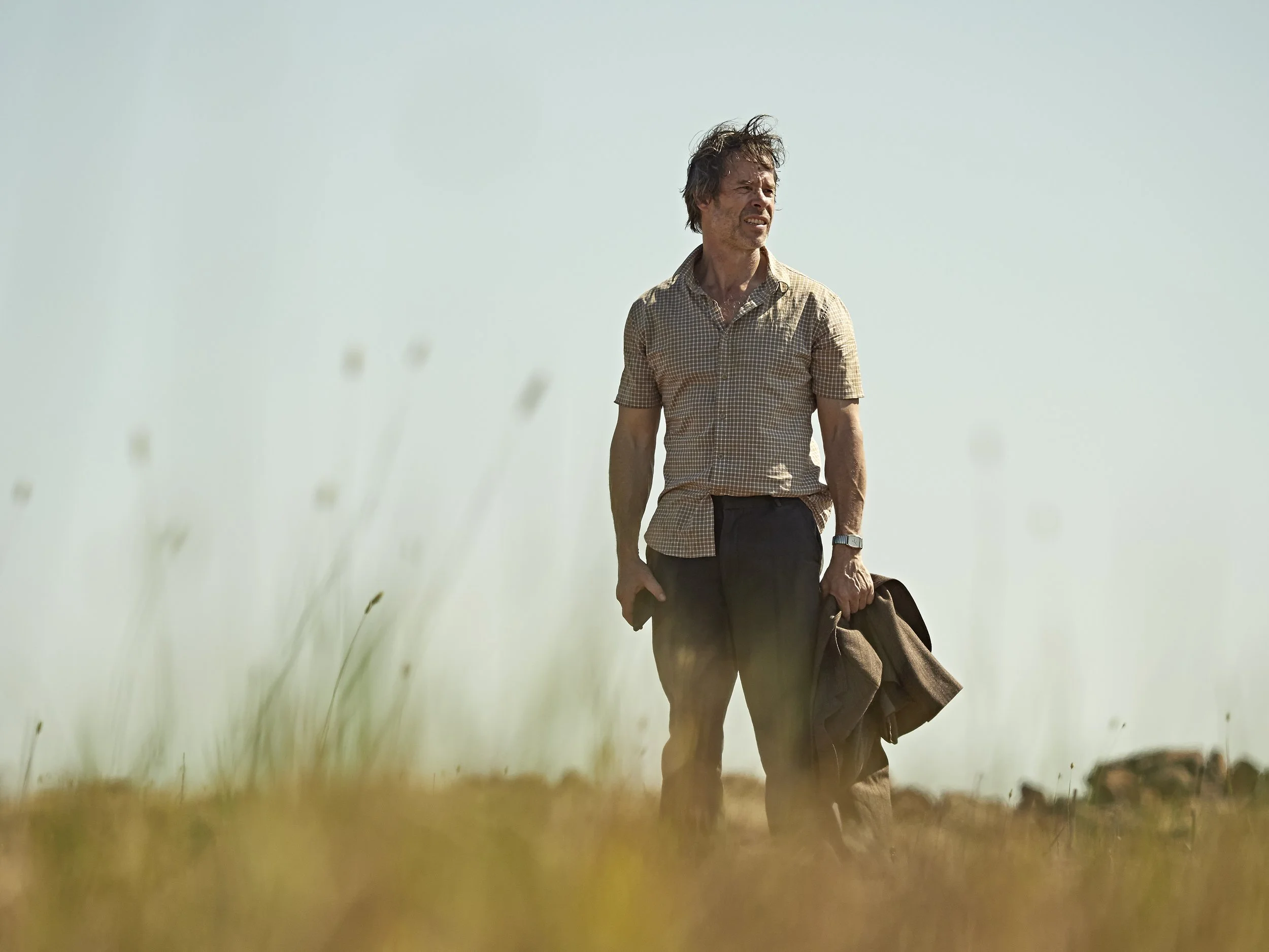 A man walking through a grassy field under a clear sky, carrying a jacket and looking into the distance.