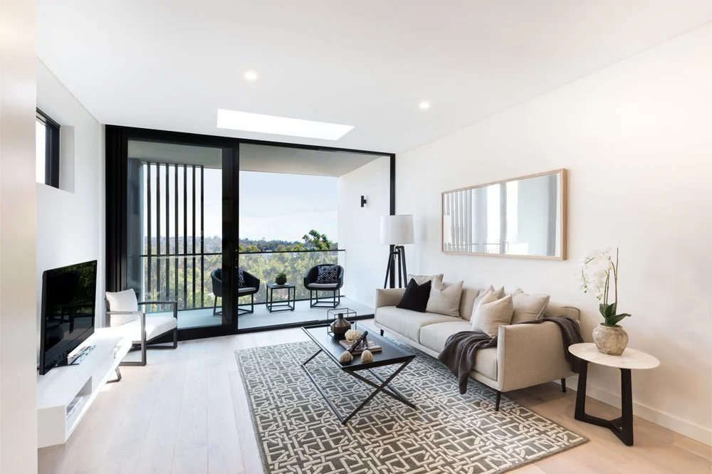 Modern living room with a gray sofa, black accent pillows, and a white armchair. Features a glass-top coffee table, patterned area rug, and a sliding glass door leading to a balcony with outdoor chairs and a view of trees.