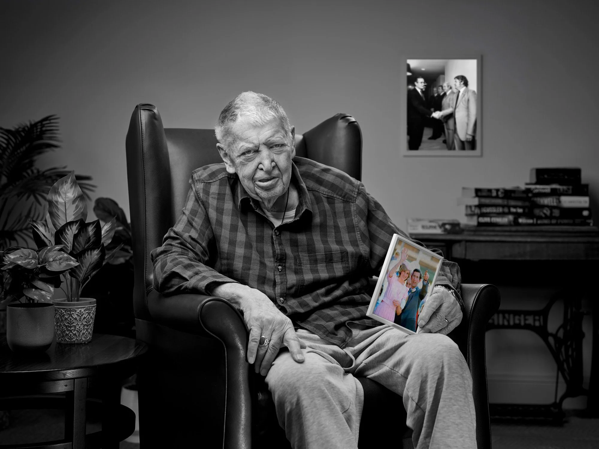 Older man sitting in a chair, holding a framed photo, with plants and a stack of books behind him, and a framed picture on the wall in the background.