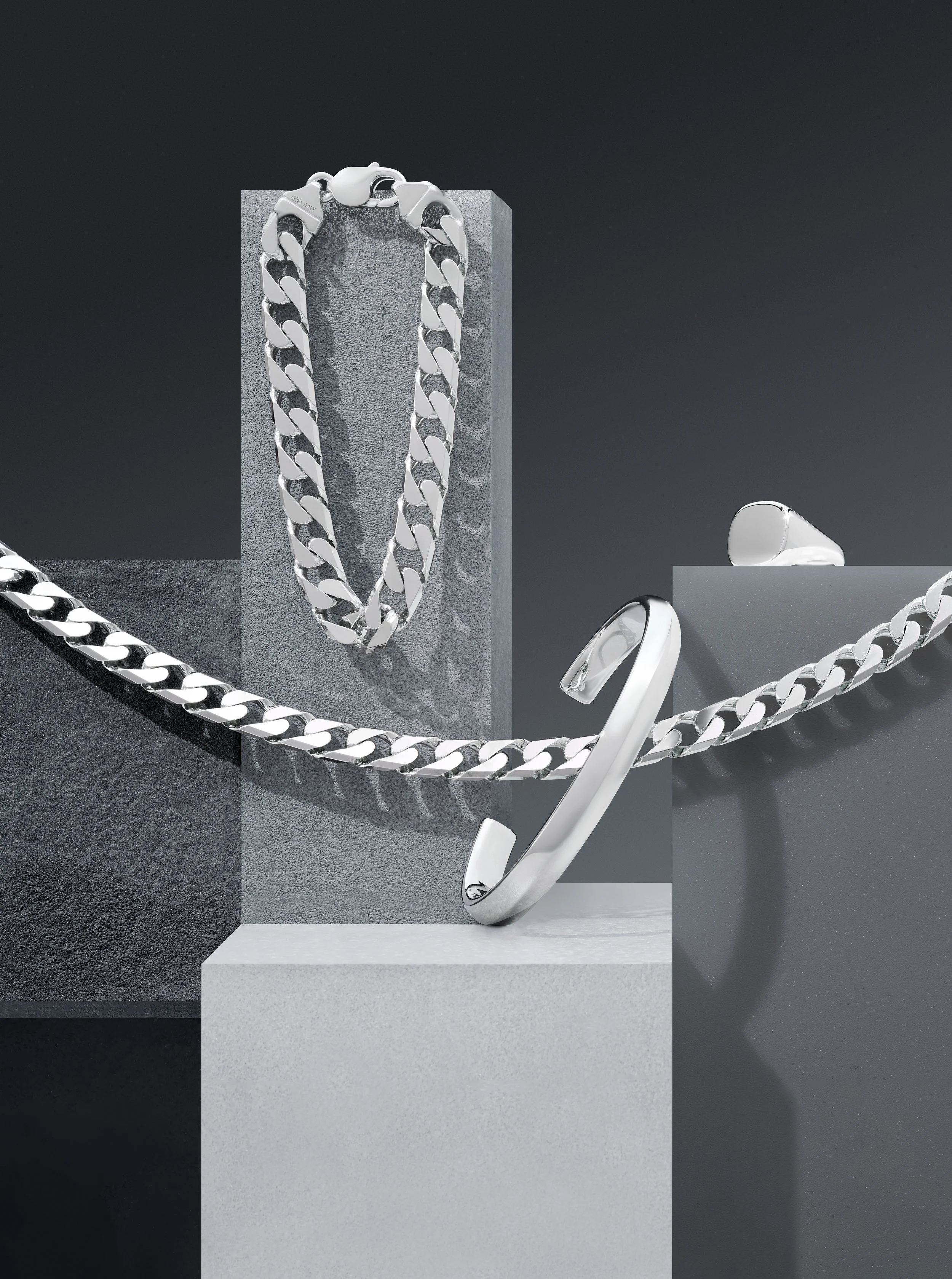 A display of silver jewelry including a chain necklace, bracelet, a ring, and a ring box on geometric gray platforms.