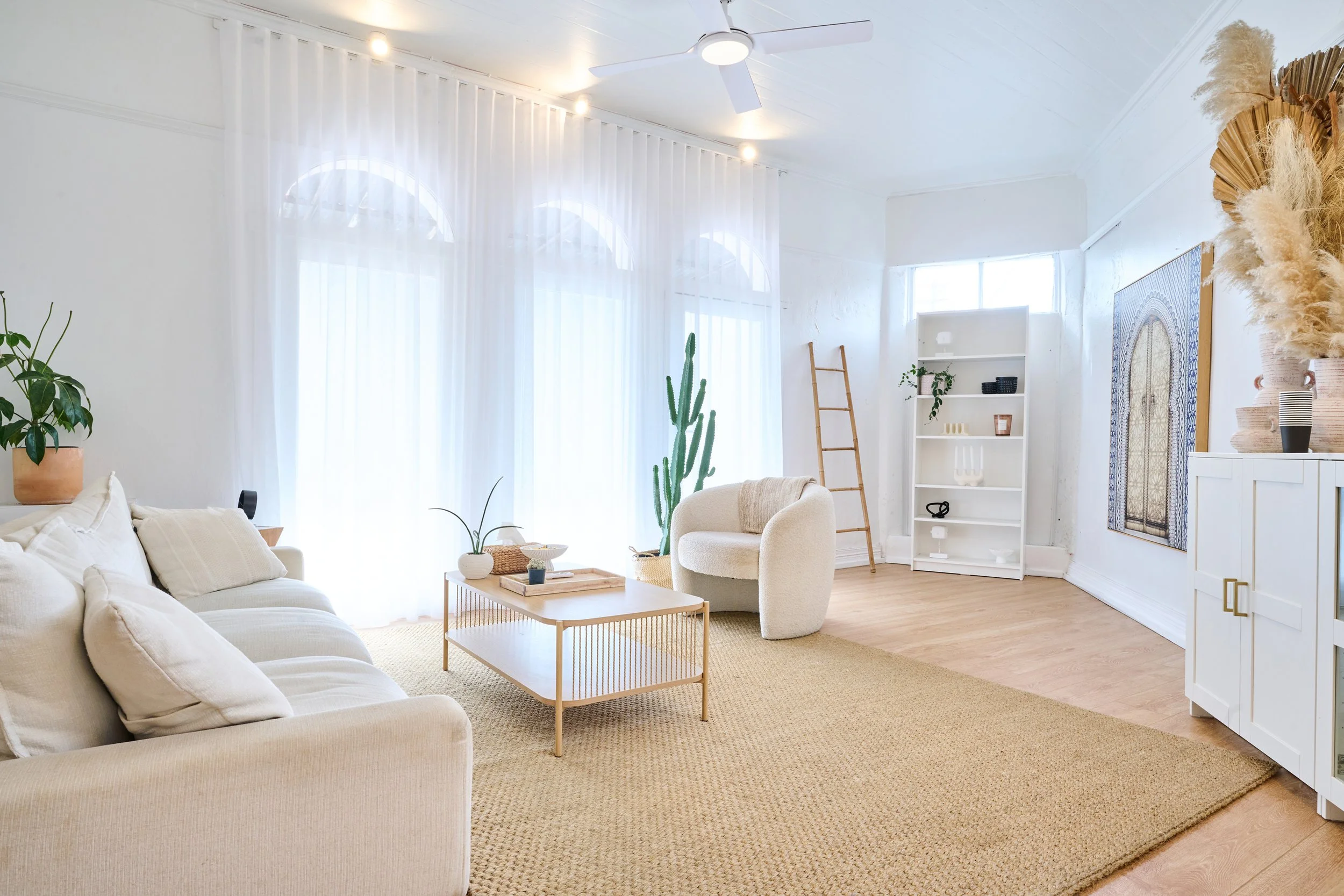 Bright living room with white walls, sheer curtains, beige sofa with pillows, wooden coffee table, white armchair, cactus plant, white shelving unit, ladder, and decorative vases with dried foliage.