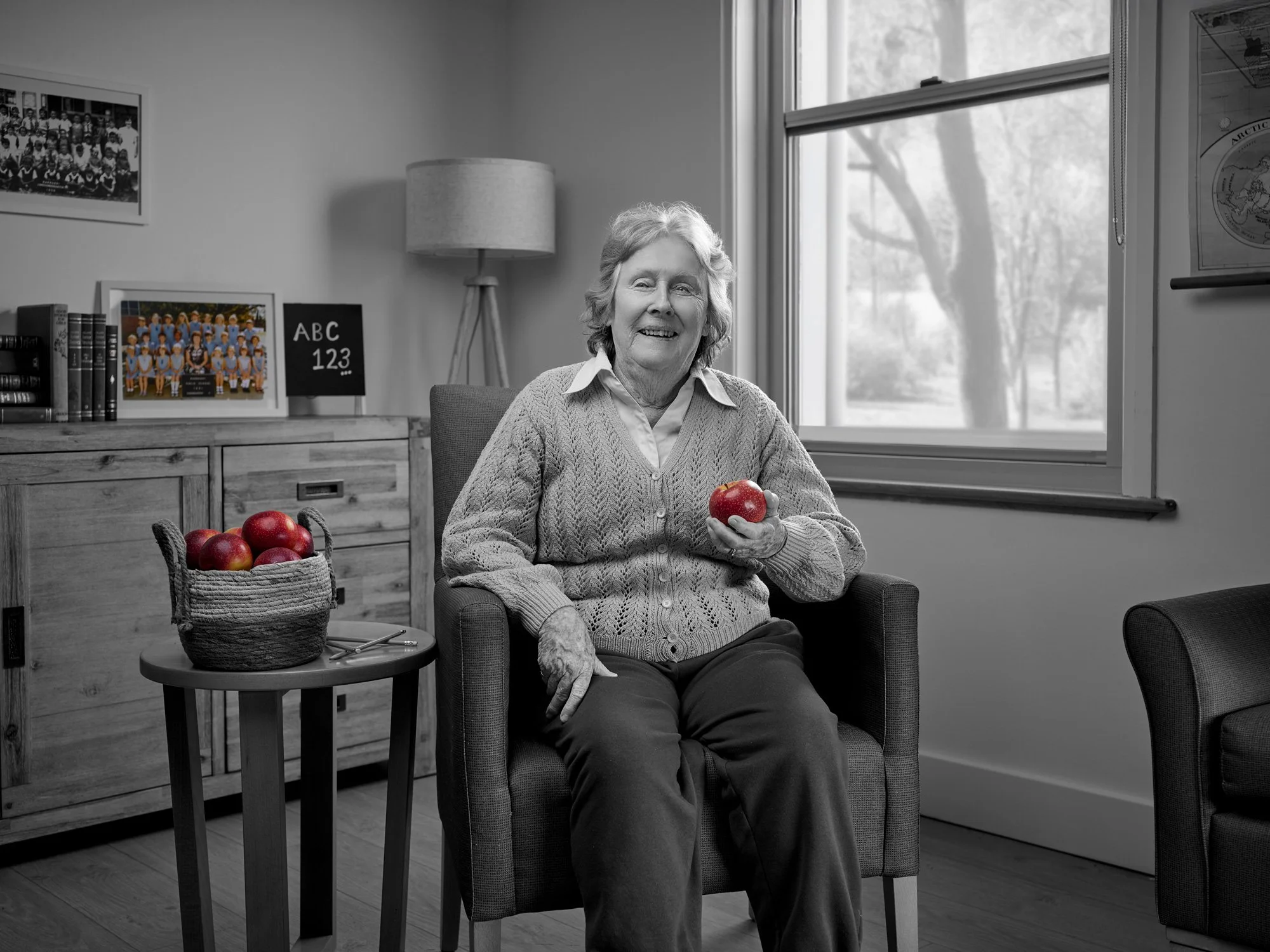 A smiling elderly woman with gray hair, wearing a cable-knit sweater over a collared shirt, sitting in a comfortable armchair in a well-lit room, holding a red apple in her right hand. There is a basket of red apples on a small table beside her. The 