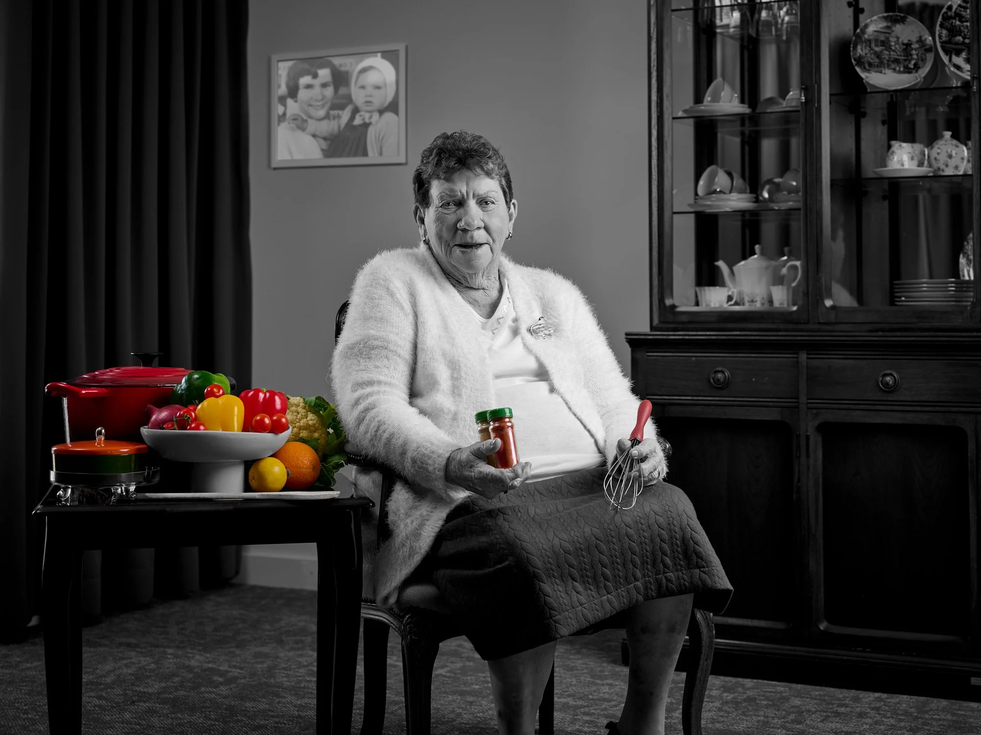 An elderly woman sitting in a dining room, holding a jar of spices and a whisk, with a bowl of colorful fruit on a table beside her. The room has a wooden cabinet with dishes and a framed photo on the wall.
