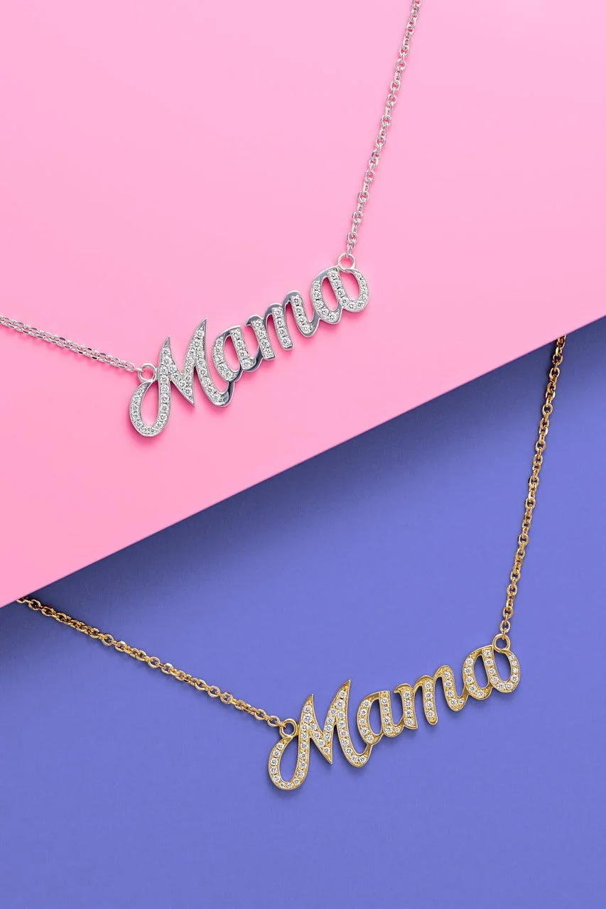 Two necklaces with the word "Mama" in cursive, one silver with clear stones on a pink background and one gold with clear stones on a blue background.