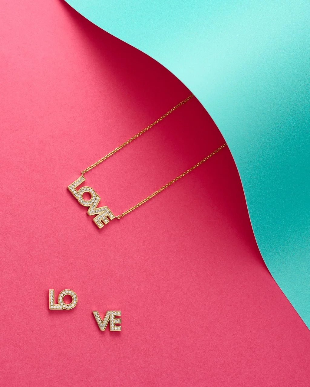 Gold necklace with 'LOVE' pendant decorated with small diamonds, lying on pink paper with a folded turquoise paper corner.