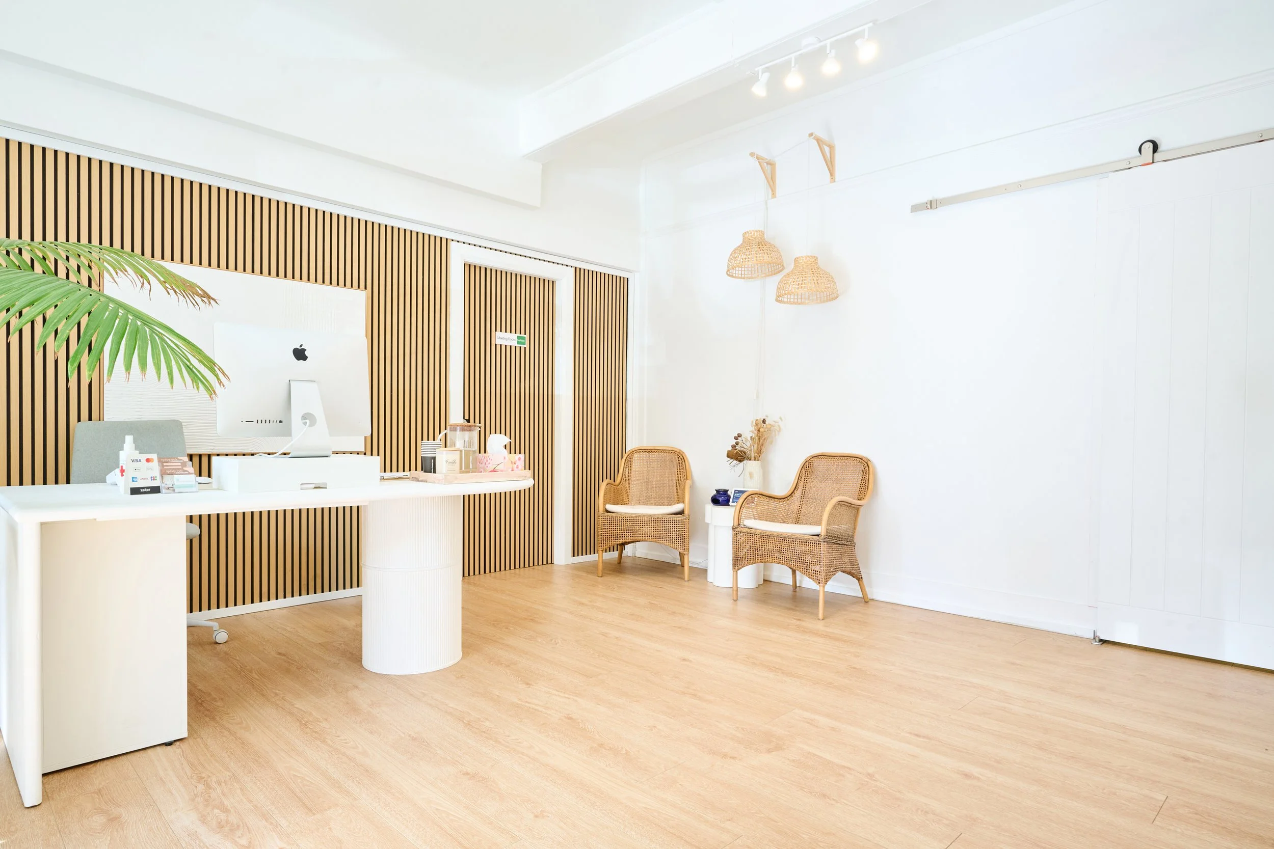 Modern reception area with a white desk, Apple iMac computer, wicker chairs, and minimal decor, featuring a wood-paneled accent wall and hanging rattan lamps.