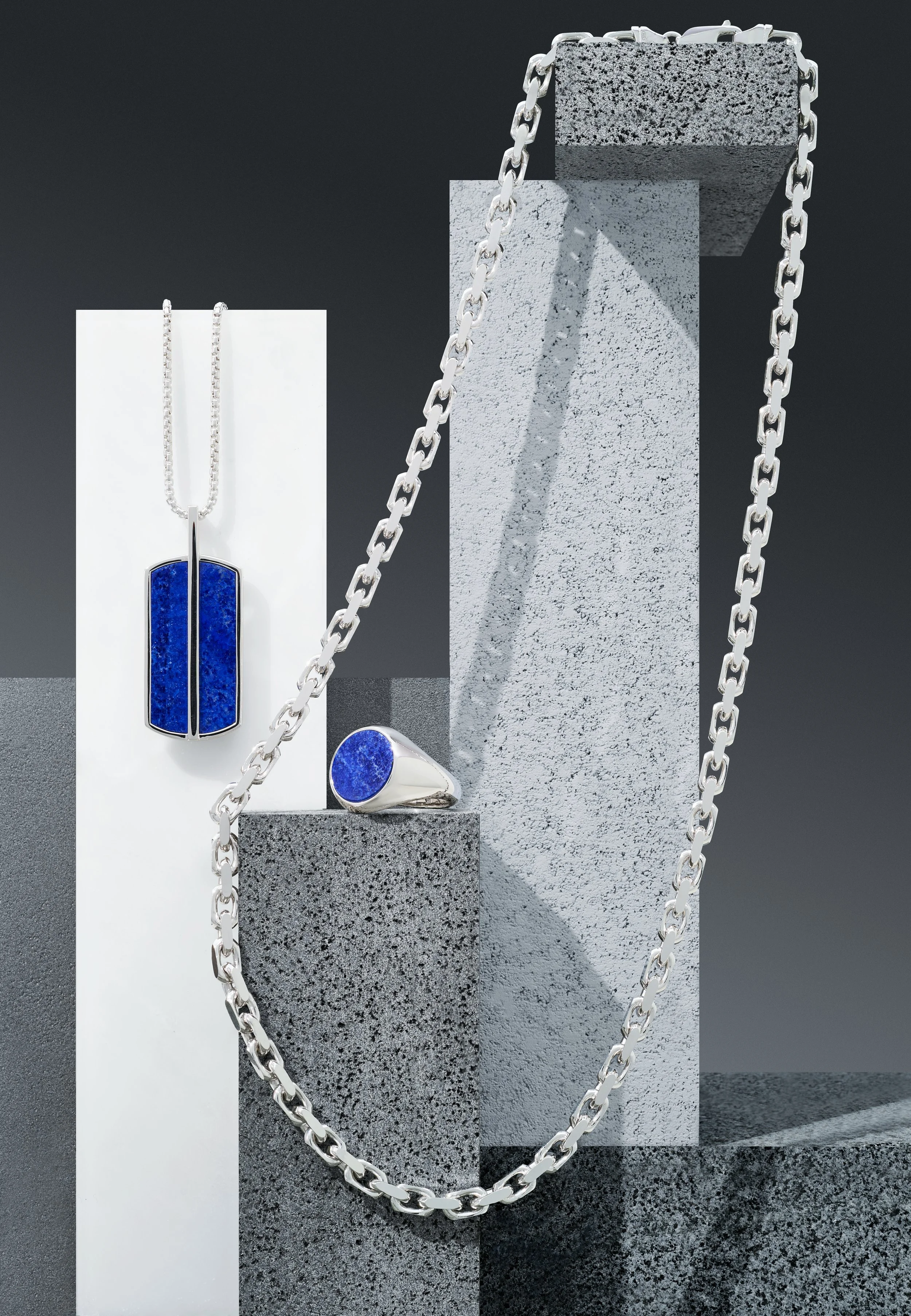 Set of silver jewelry with deep blue stone accents, including a pendant necklace, a ring, and a chain, displayed on textured gray blocks.