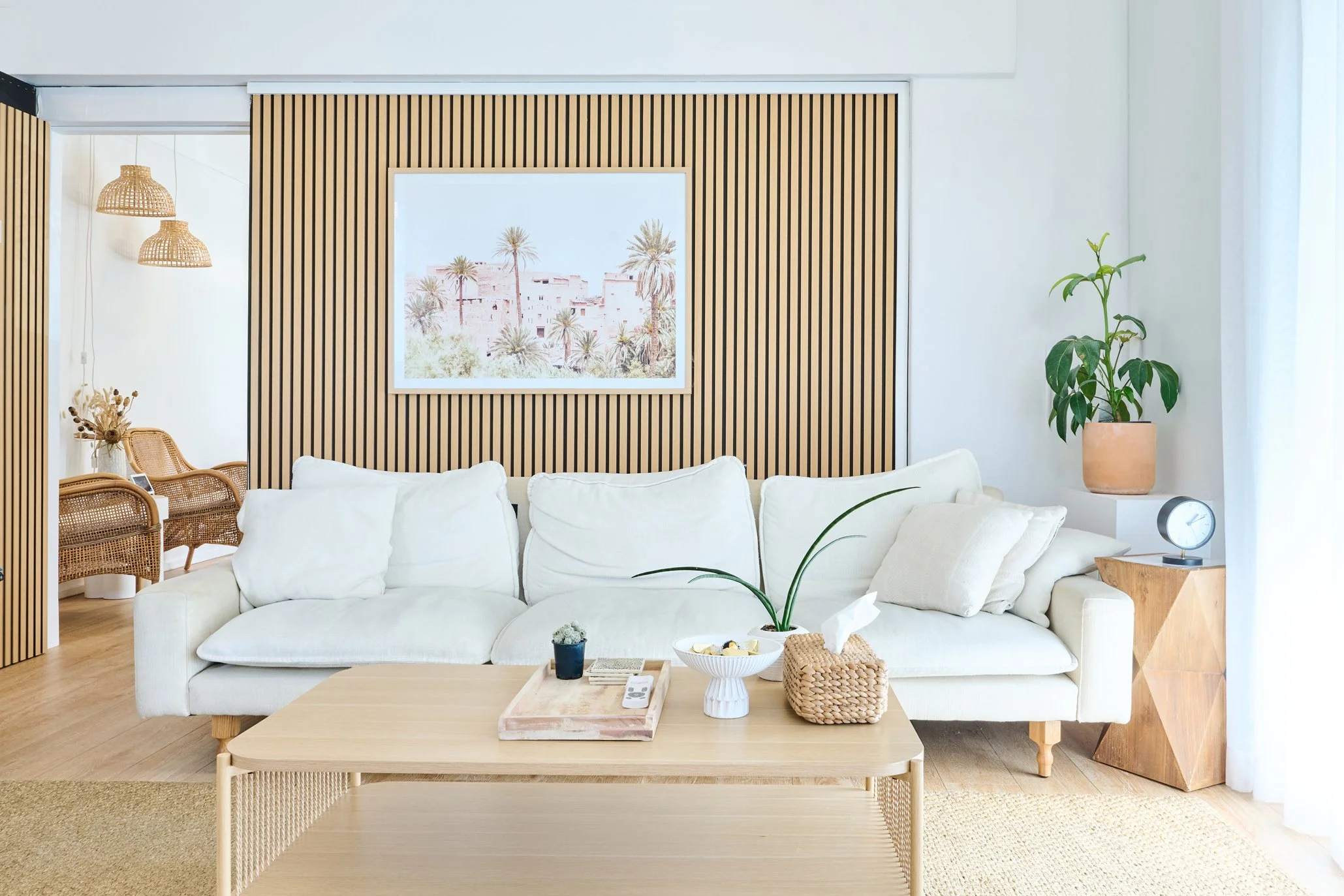 Living room with a white sofa, a light wooden coffee table, a large plant on a side table, and wall art of a desert scene with palm trees. Rattan chairs and pendant lights are visible in the background.