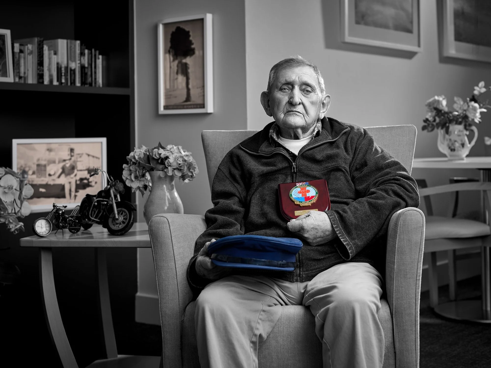 An elderly man sitting in a beige armchair in a living room, holding a framed badge or medal, with a blue pouch on his lap. The room has framed pictures on the wall, a bookshelf with books, and a side table with decorative items and flowers.