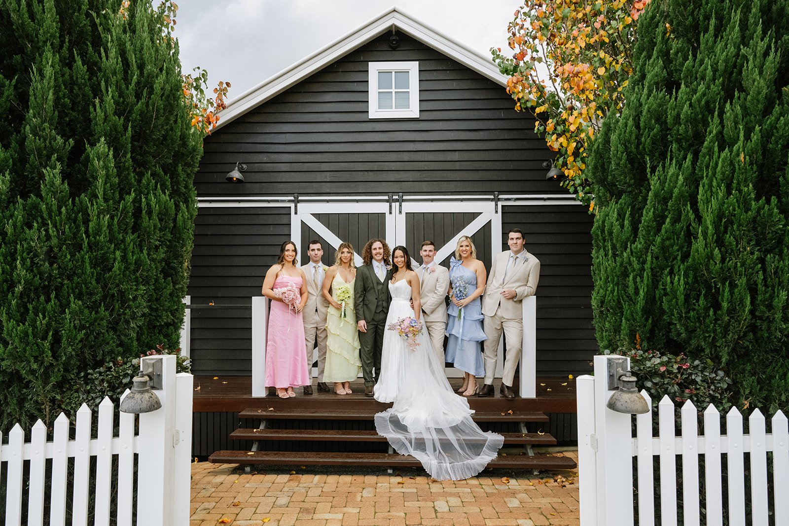 A wedding party stand on the steps of Black Hall Kalbar with their friends. The dresses are in mis matched pastel colours and the guys are wearing sage suits