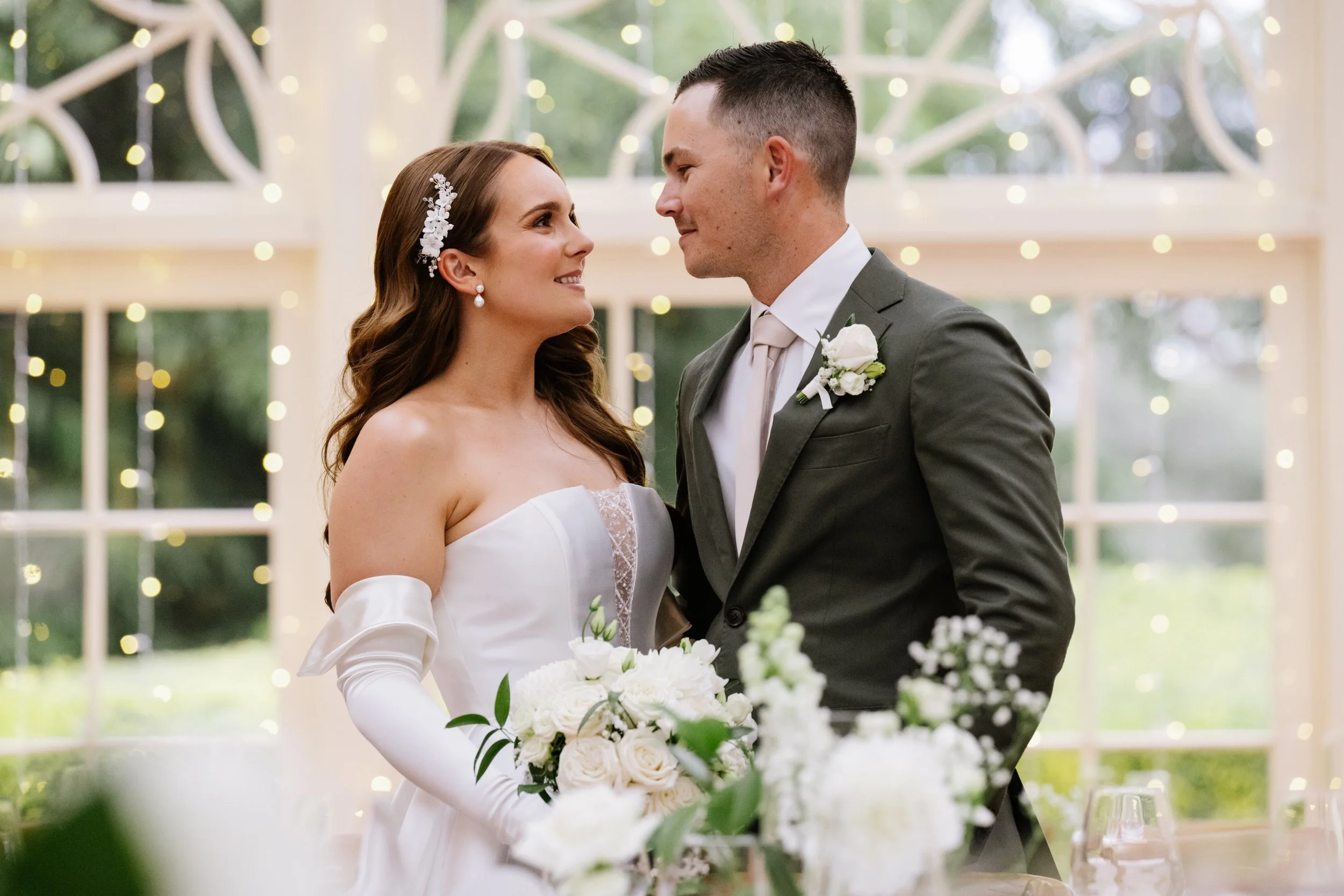 A you couple gazing in to each others eyes in the historic gabbinbar  in Toowoomba . The woman is wear a classic white wedding dress and the man in a deep sage suit . The woman is wearing long white gloves and her hair is  in glam waves .