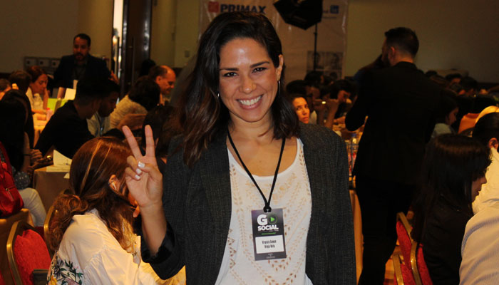 Virginia Saman sonriendo, haciendo señal de paz con dos dedos en un evento con muchas personas en el fondo.