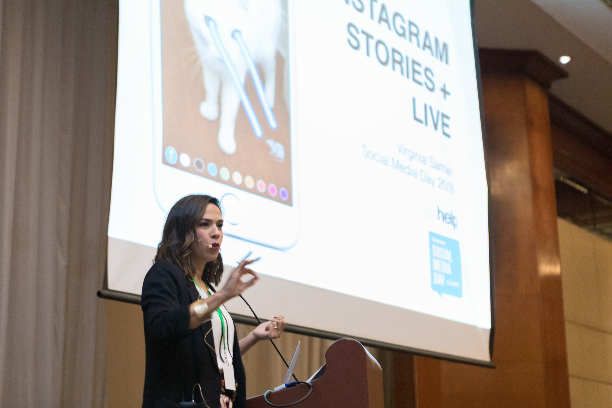 Virginia Saman hablando en una conferencia frente a una pantalla que muestra un póster sobre Instagram Stories y en vivo, en el evento Social Media Day 2019.