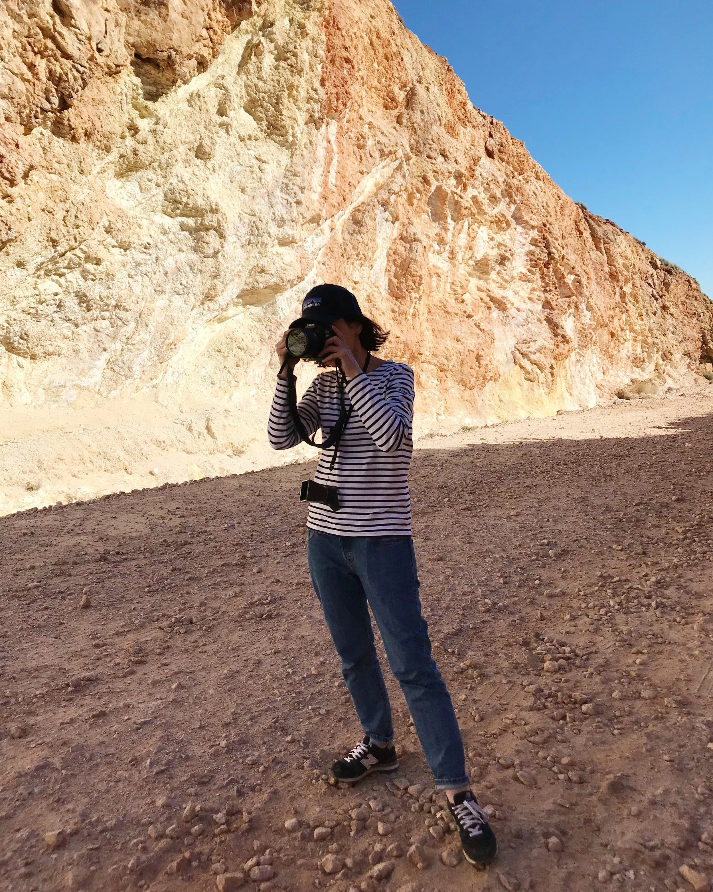 Person wearing a striped shirt, jeans, and a baseball cap taking a photo with a camera in front of a large rocky cliff under a clear blue sky.