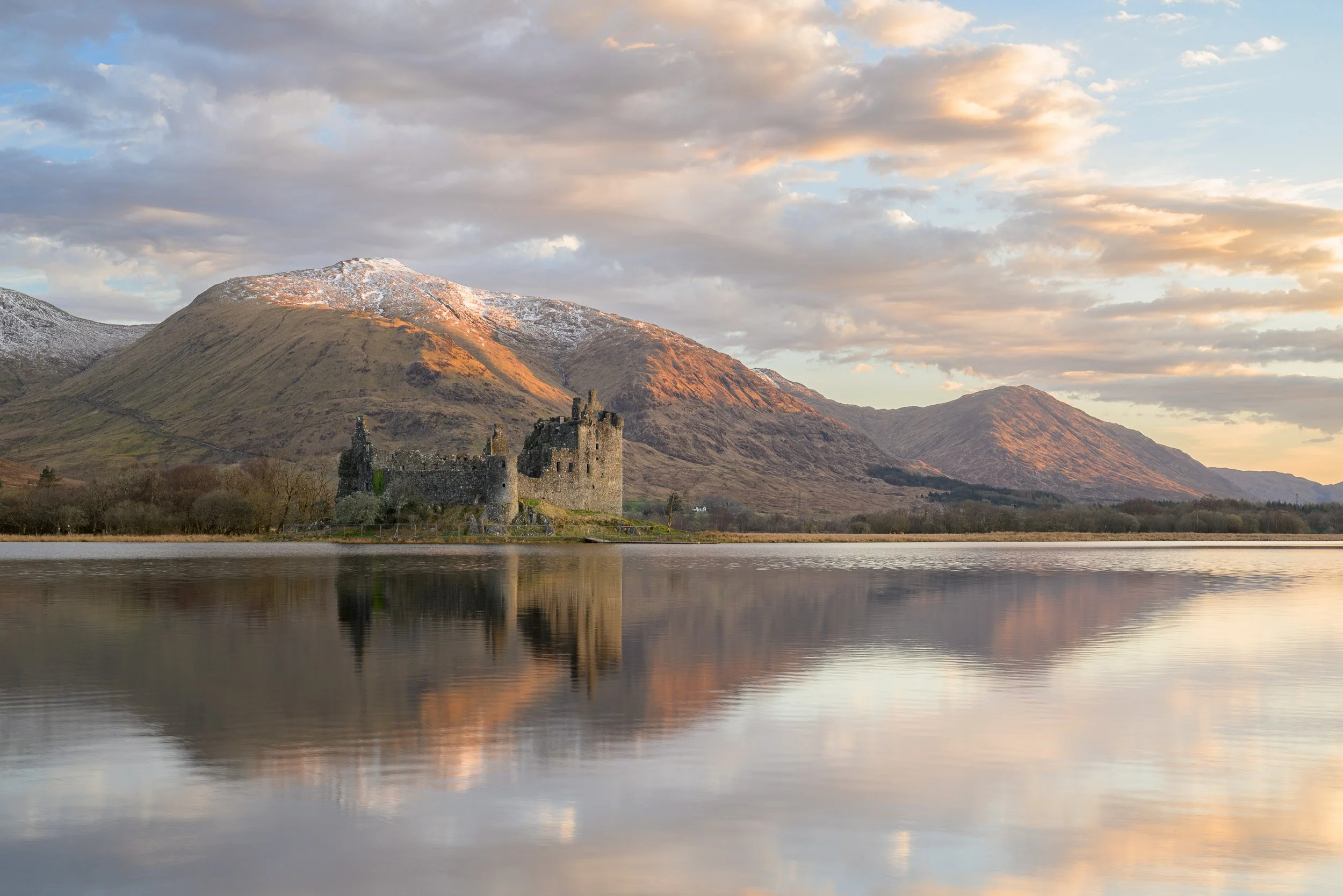 A sunrise photograph of Kilchurn Castle on Loch Awe with snow on the mountains and a reflection on the loch with a beautiful sky.