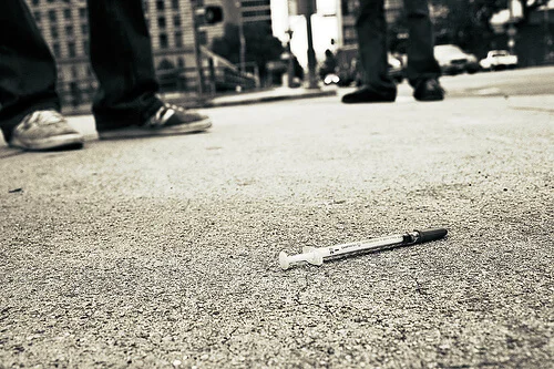 A used syringe that was possibly filled with illegal drugs laying on the street near some people.