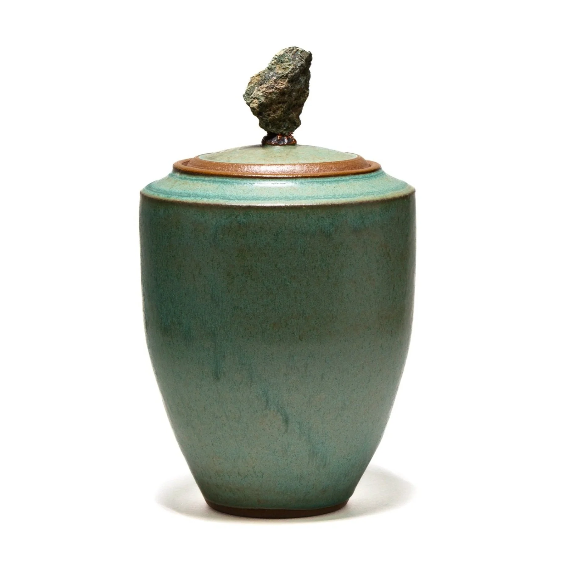 A ceramic vase with a rounded bodice, tapering toward the base, glazed in a greenish hue, with a small, rough, dark-colored rock balanced on top of the lid.