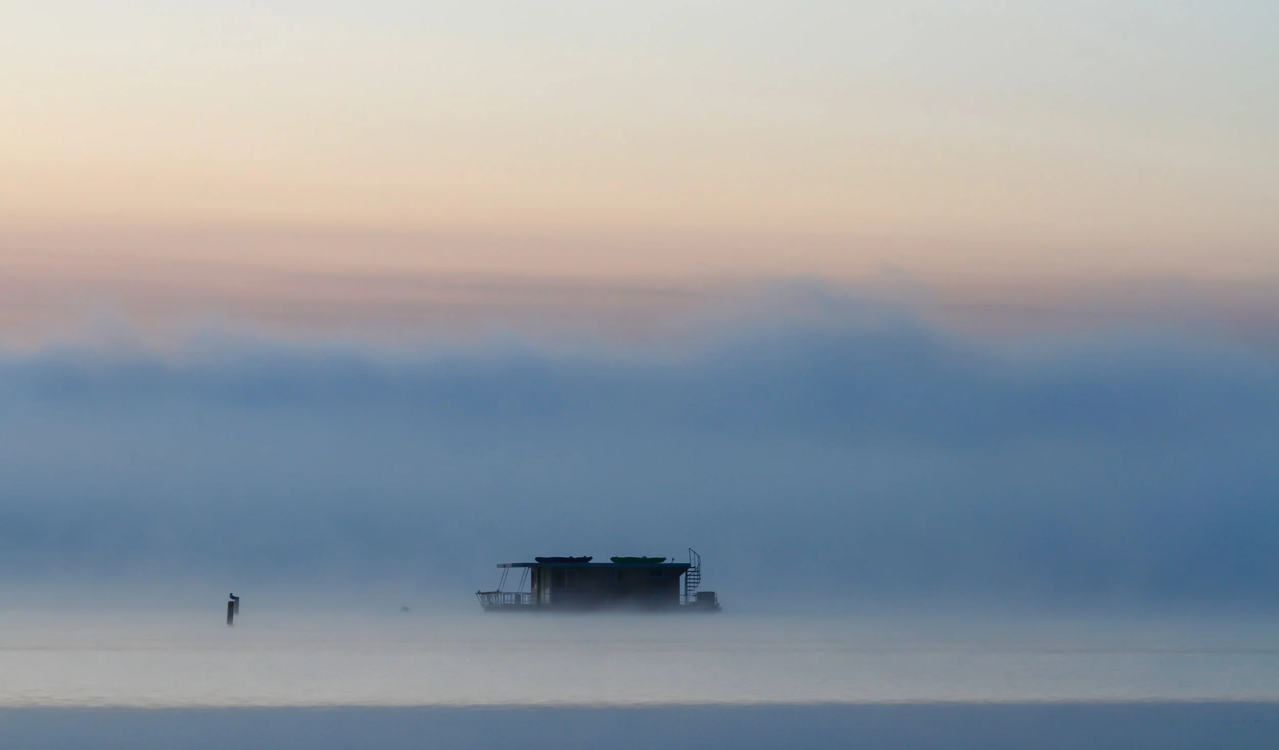A boat floating on a foggy body of water during twilight with a pastel-colored sky.