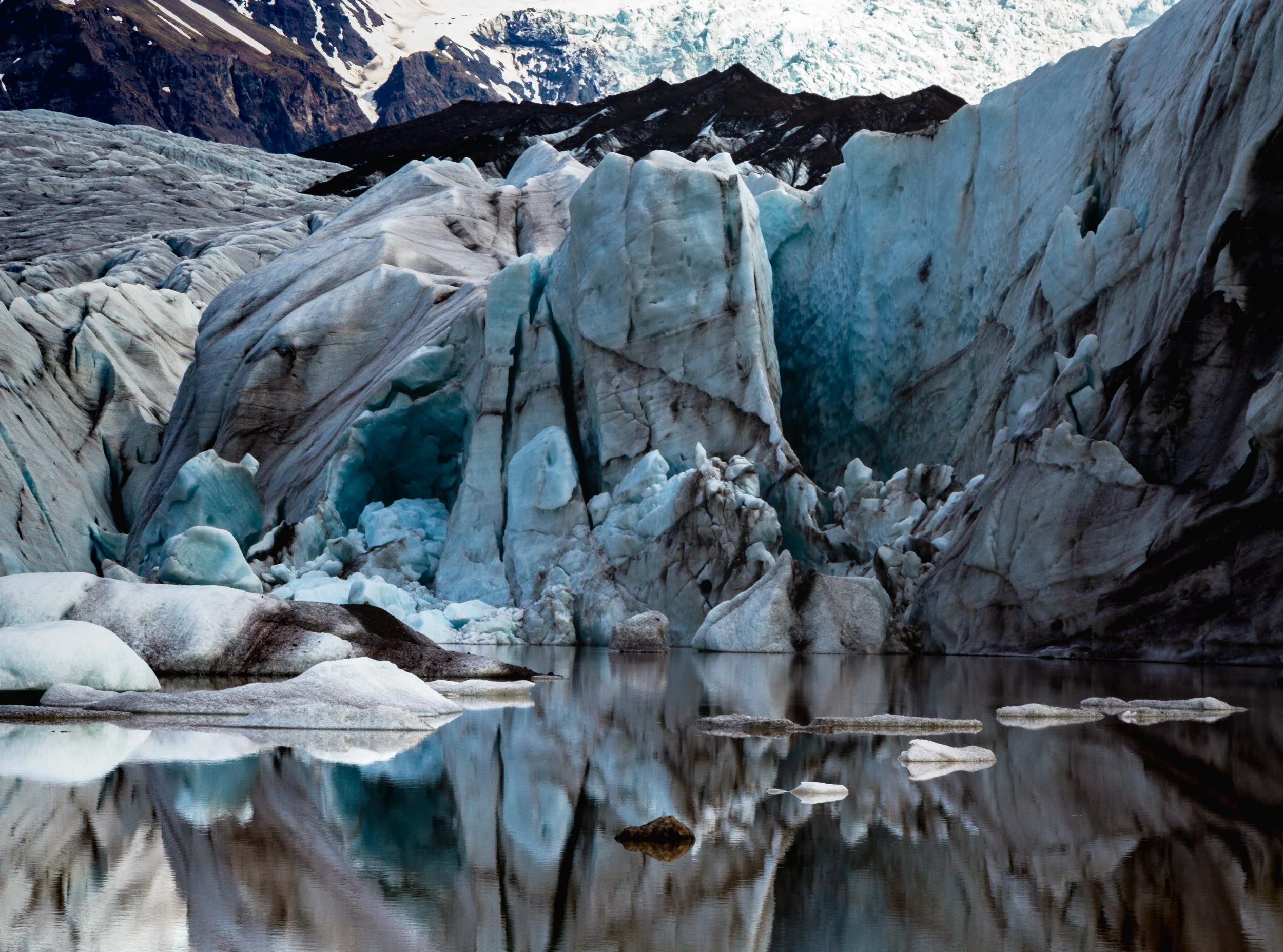 Glacier ice with blue crevasses and a reflection in a body of water, surrounded by mountains.