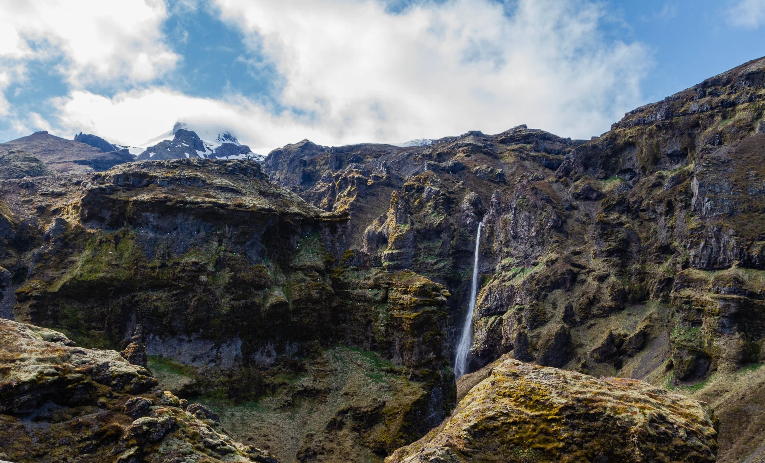 A rugged mountain landscape with a small waterfall cascading down rocky terrain and snow-capped peaks under a partly cloudy sky.