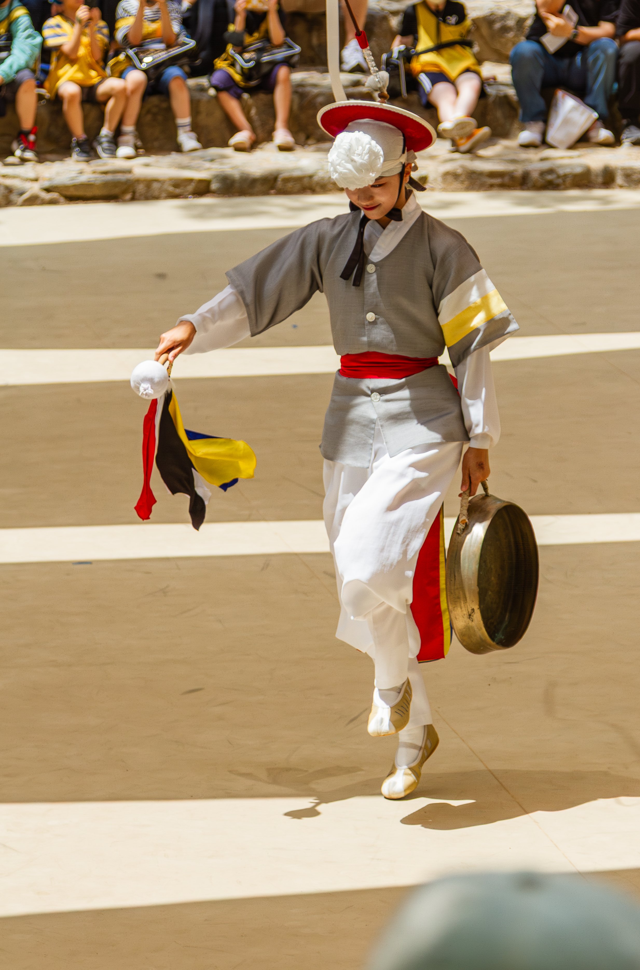 A performer dressed in traditional Korean attire, holding a gong and a small flag, performing a dance or ritual. In the background, children are seated outdoors, watching the performance.