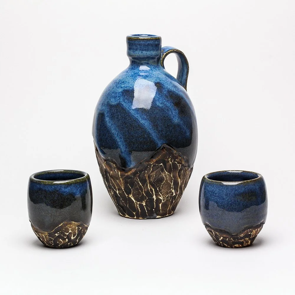 Ceramic pitcher and two cups with blue and black glaze and earthy brown textured bands at the bottom.