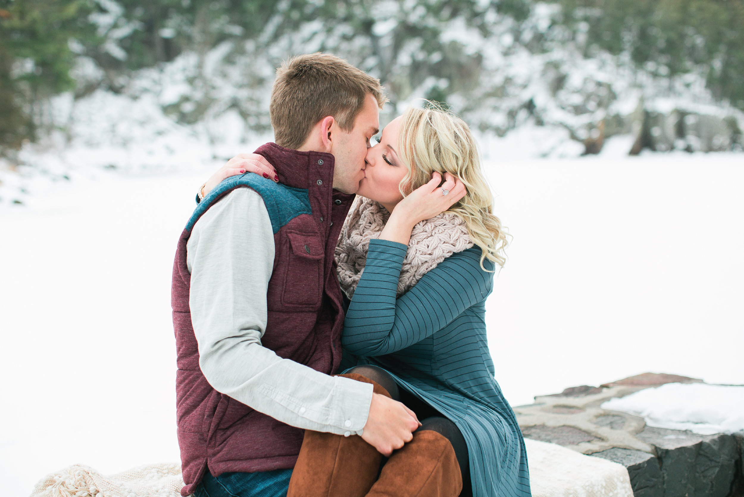 Taylors Falls Minnesota snowy winter engagement session couple sitting and kissing with woods background