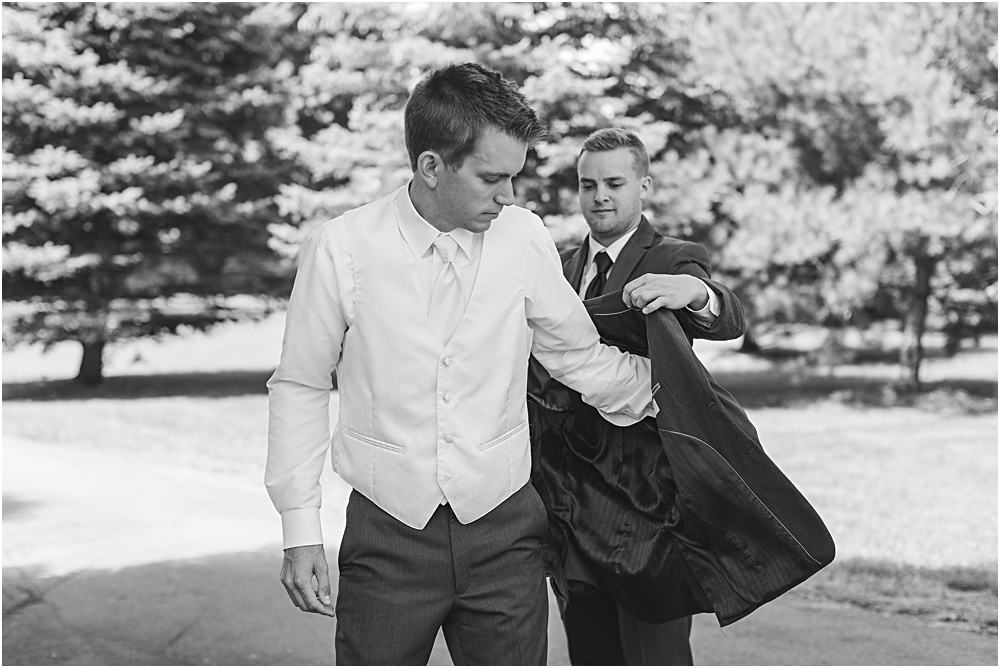 Groom getting ready for Minnesota summer wedding day in dark gray suit and white vest best man helping groom into suit