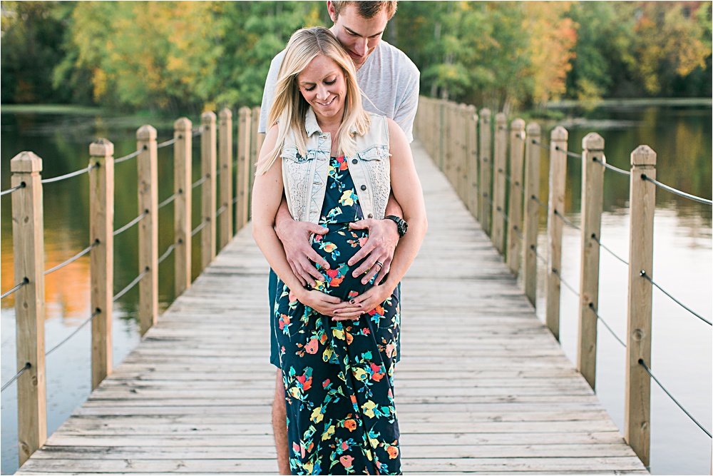 Husband and wife on bridge for fall Minnesota maternity photos showing bump with floral maxi dress and denim vest