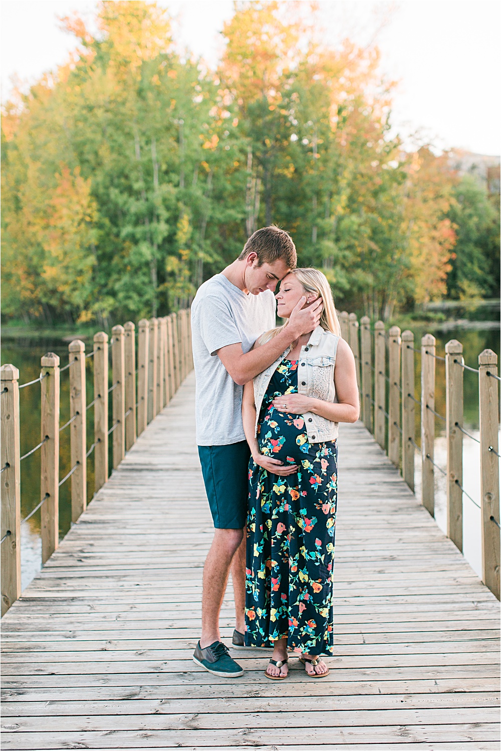 Husband and wife on bridge for fall Minnesota maternity photos showing bump with floral maxi dress and denim vest