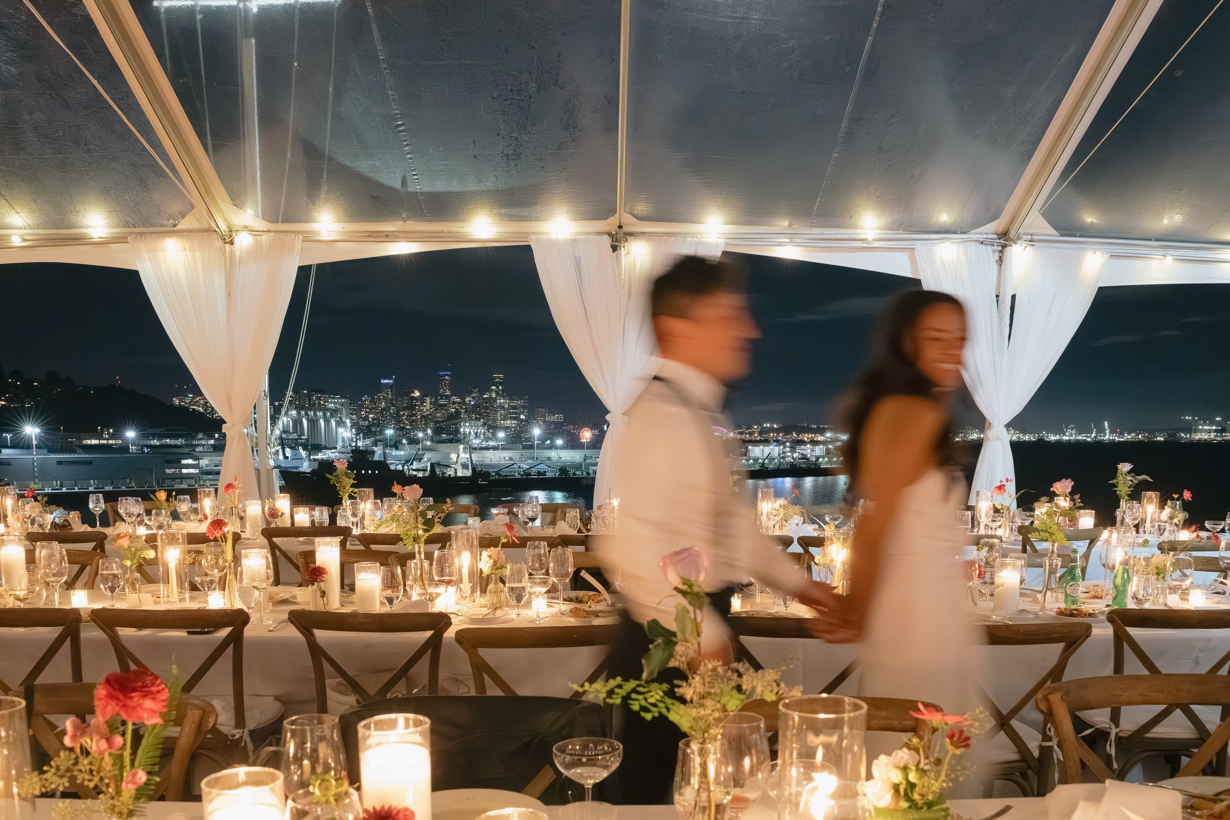 Reception table setting with a romantic warm glow from numerous white pillar candles, the beautiful seattle skyline, and pops of colorful florals.