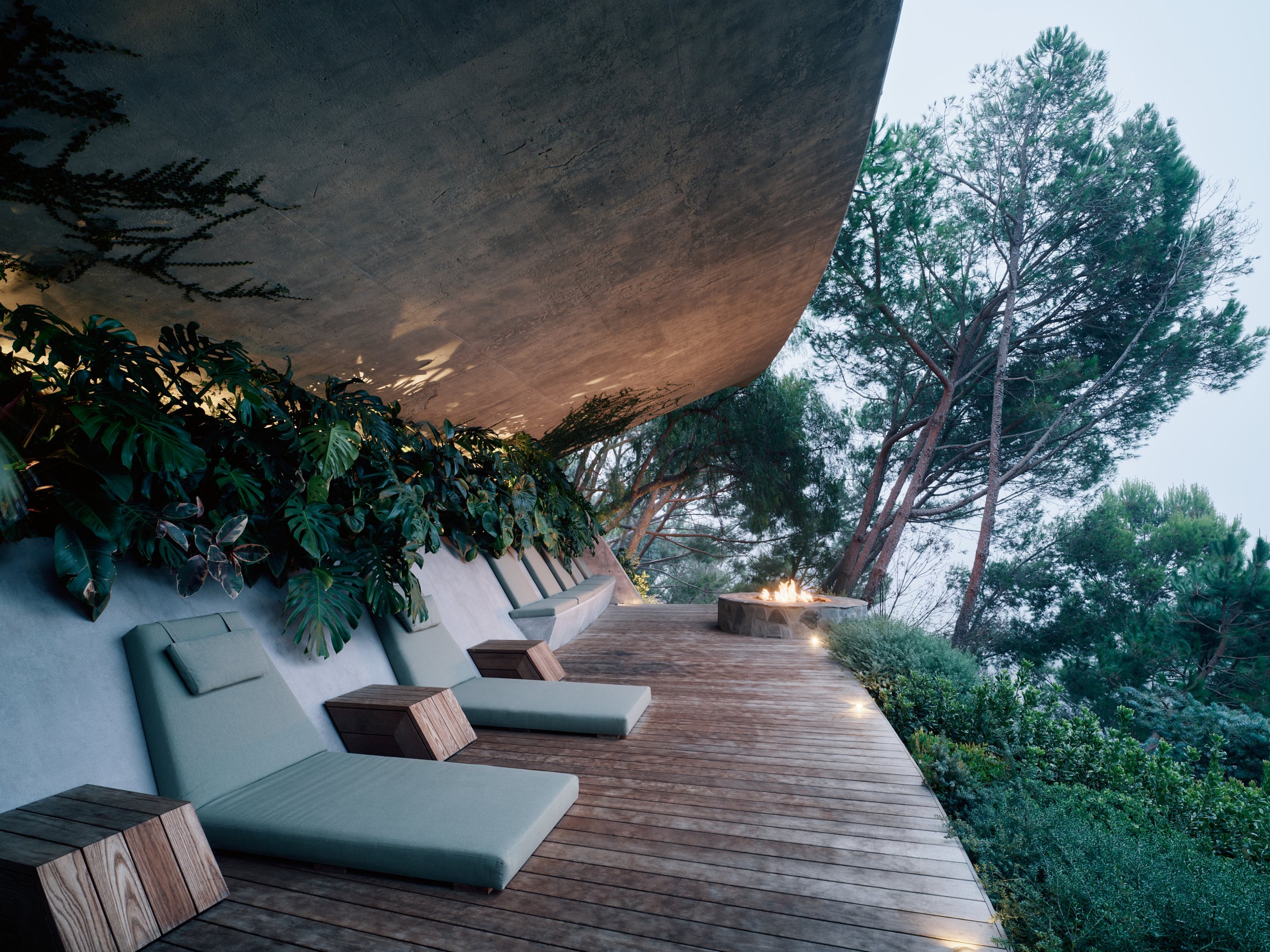 Evening view of the new fire pit deck at Lautner’s Castle by Conner + Perry Architects. The shot shows the built-in lounge furniture, lush tropical plants like Monstera, and a stone fire pit alit under the original 1982 concrete pool cantilever.