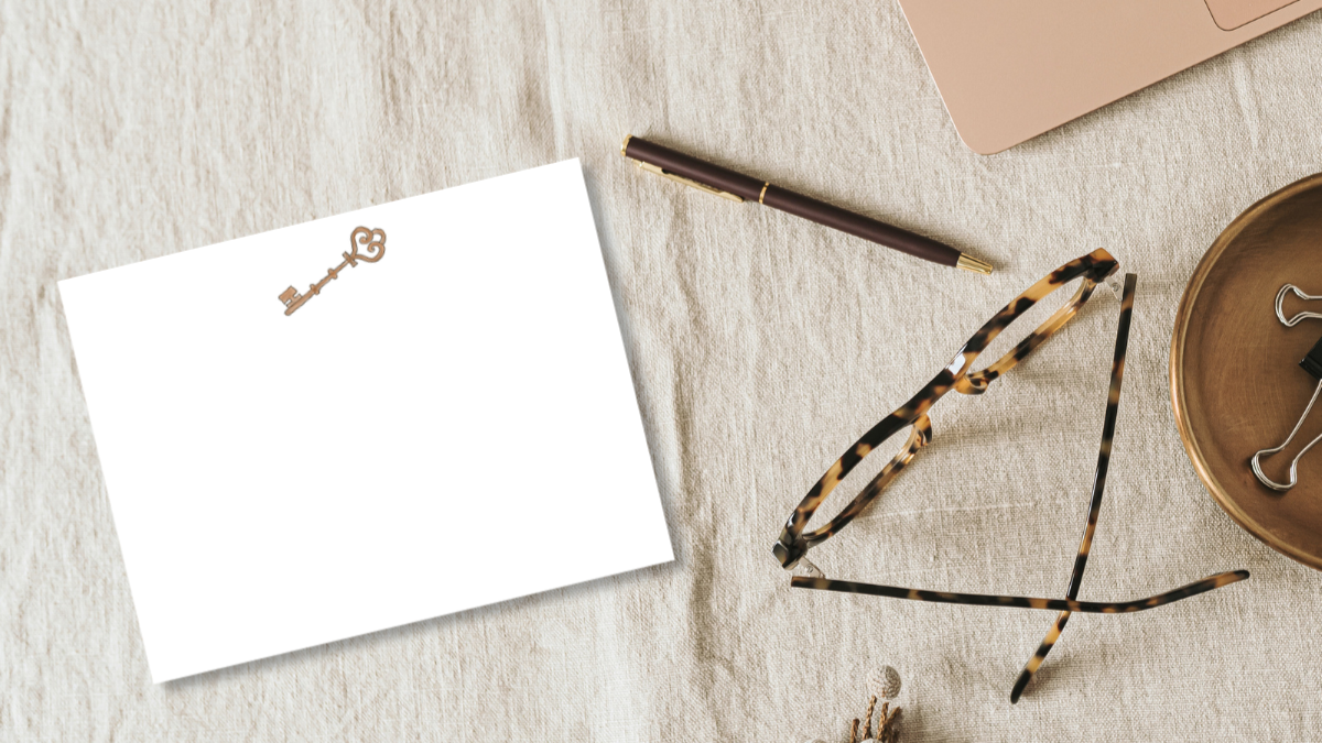 birds eye view of an office desk with a laptop, pen, eye glasses, and a note card with an illustration of an antique door key designed by Viola and Beatrice Stationers