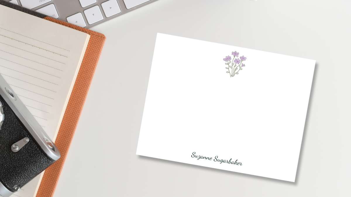 office desk with a keyboard, notebook, camera, and note card with an illustrated bunch of flowers and the name suzanne sugarbaker