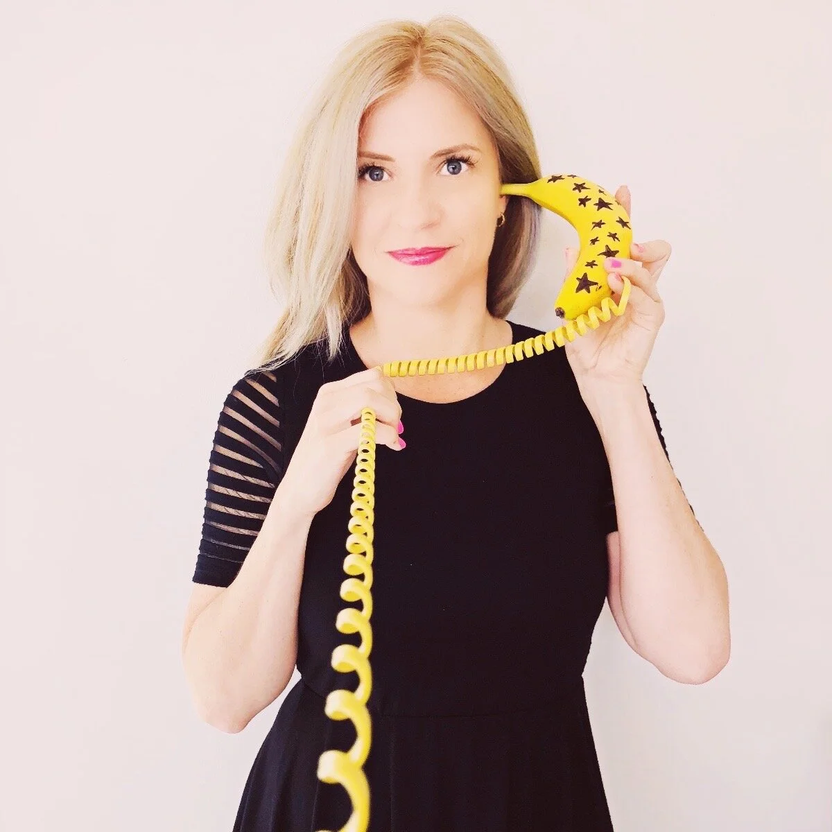 You can call the Style Rituals hotline! This is an image of Colleen McCann of Style Rituals standing in a black tea dress holding a bright yellow banana phone.