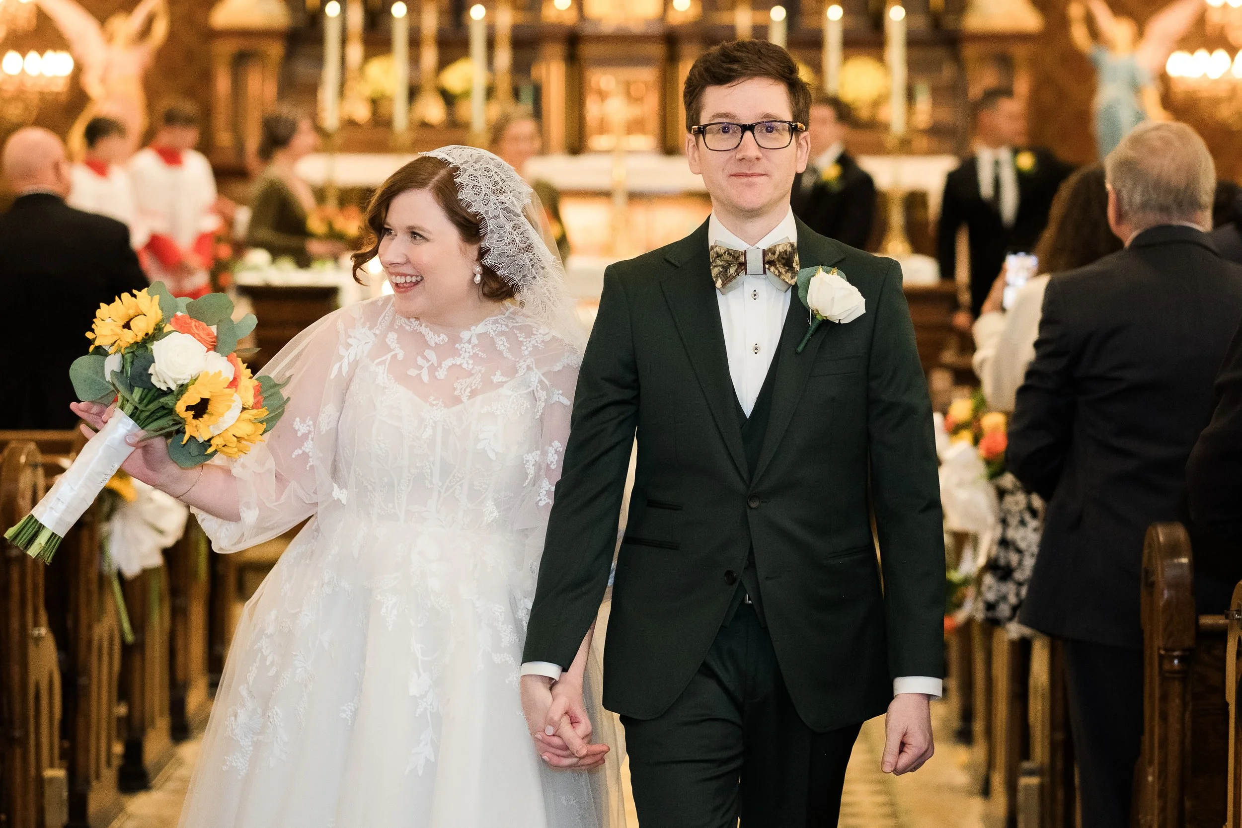 Bride and groom walking down a church aisle after their wedding ceremony, wearing a lace wedding dress and dark green suit with sage green brown plum bowtie knotzland bow tie.