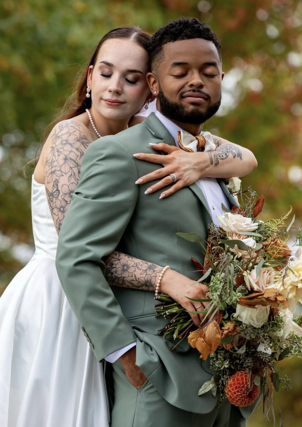 Wedding couple embracing during an outdoor fall ceremony portrait, featuring a sage green suit, patterned knotzland bow tie, and seasonal bouquet.