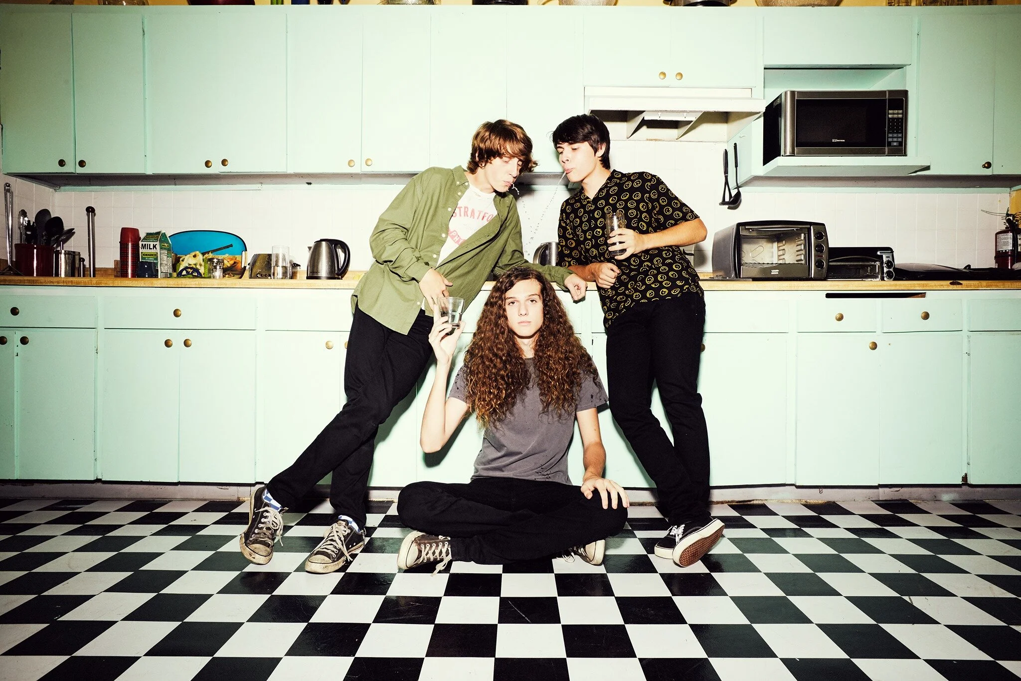 Three young adults in a kitchen with mint green cabinets and black-and-white checkered floor. Two are standing, leaning on the counter, holding drinks. One is sitting on the floor with legs crossed, also holding a drink, with curly hair and a serious