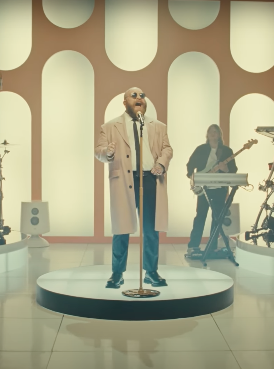 A man wearing sunglasses, a beige coat, and a black tie singing into a microphone on a circular stage, with a keyboardist playing behind him. The background has a retro-style pattern with arches in a warm color palette.