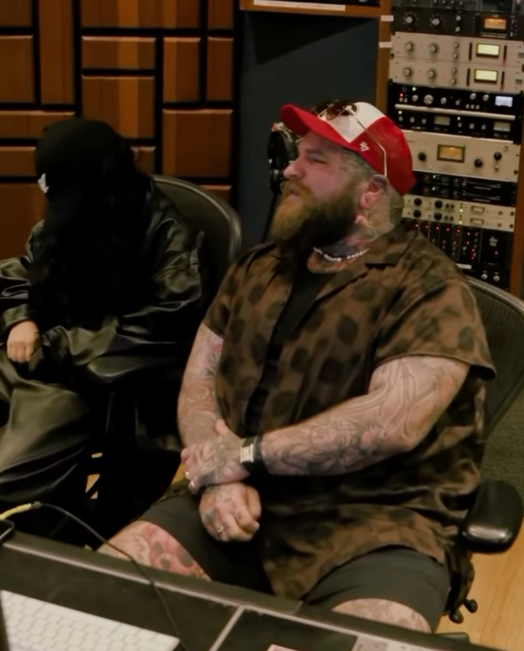 A bearded man with tattoos on his arms, wearing a brown leopard print shirt, black shorts, a red and white trucker hat, and sunglasses on his hat, sitting in a recording studio with audio equipment behind him.