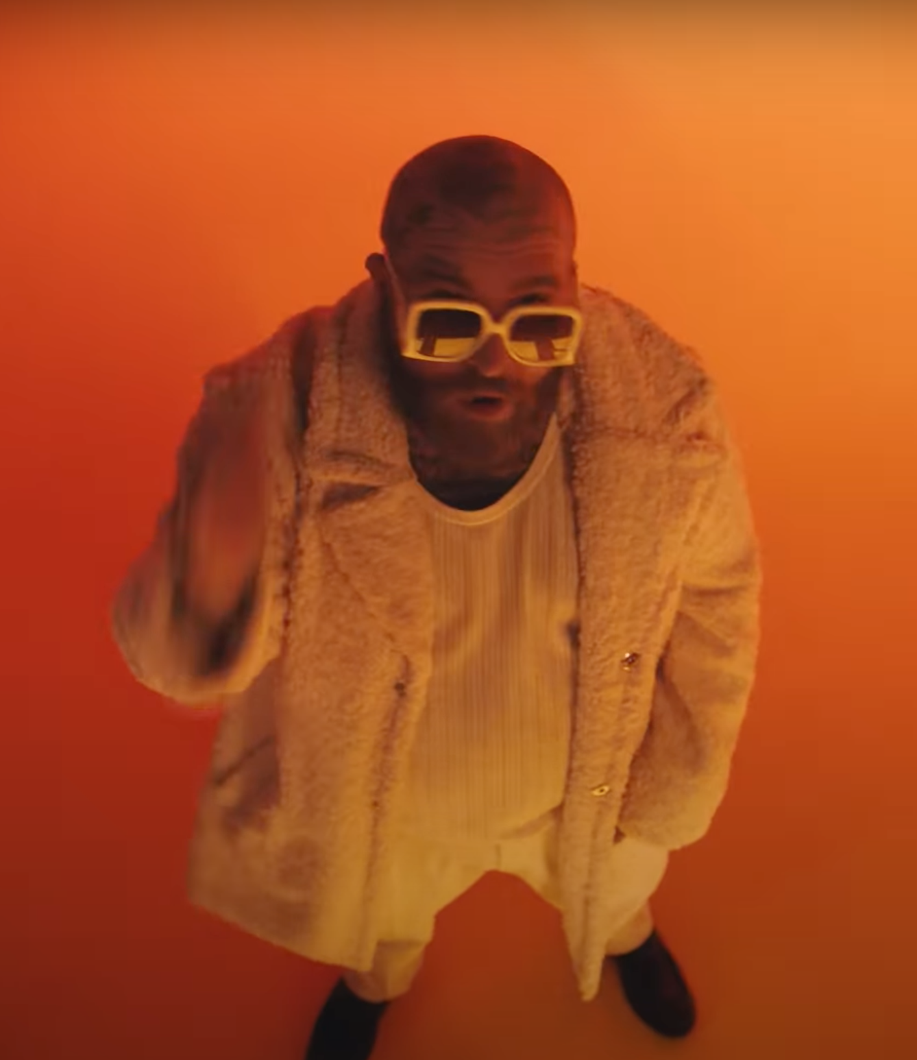 Man wearing yellow sunglasses, a beige coat, and a white shirt, standing against an orange background.