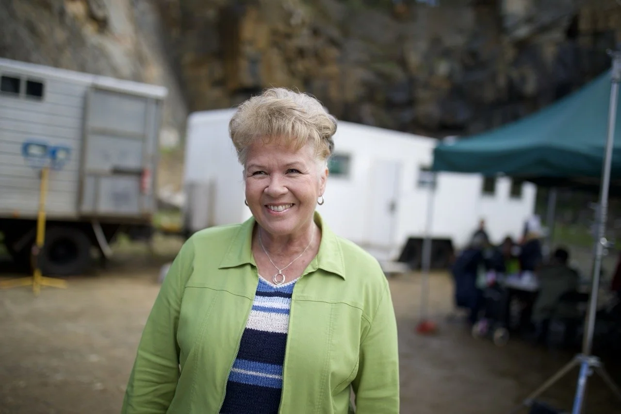 Smiling older woman in lime green jacket outdoors with trailers and a tent in the background actor Sarah Woods carol hobday koko a red dog story