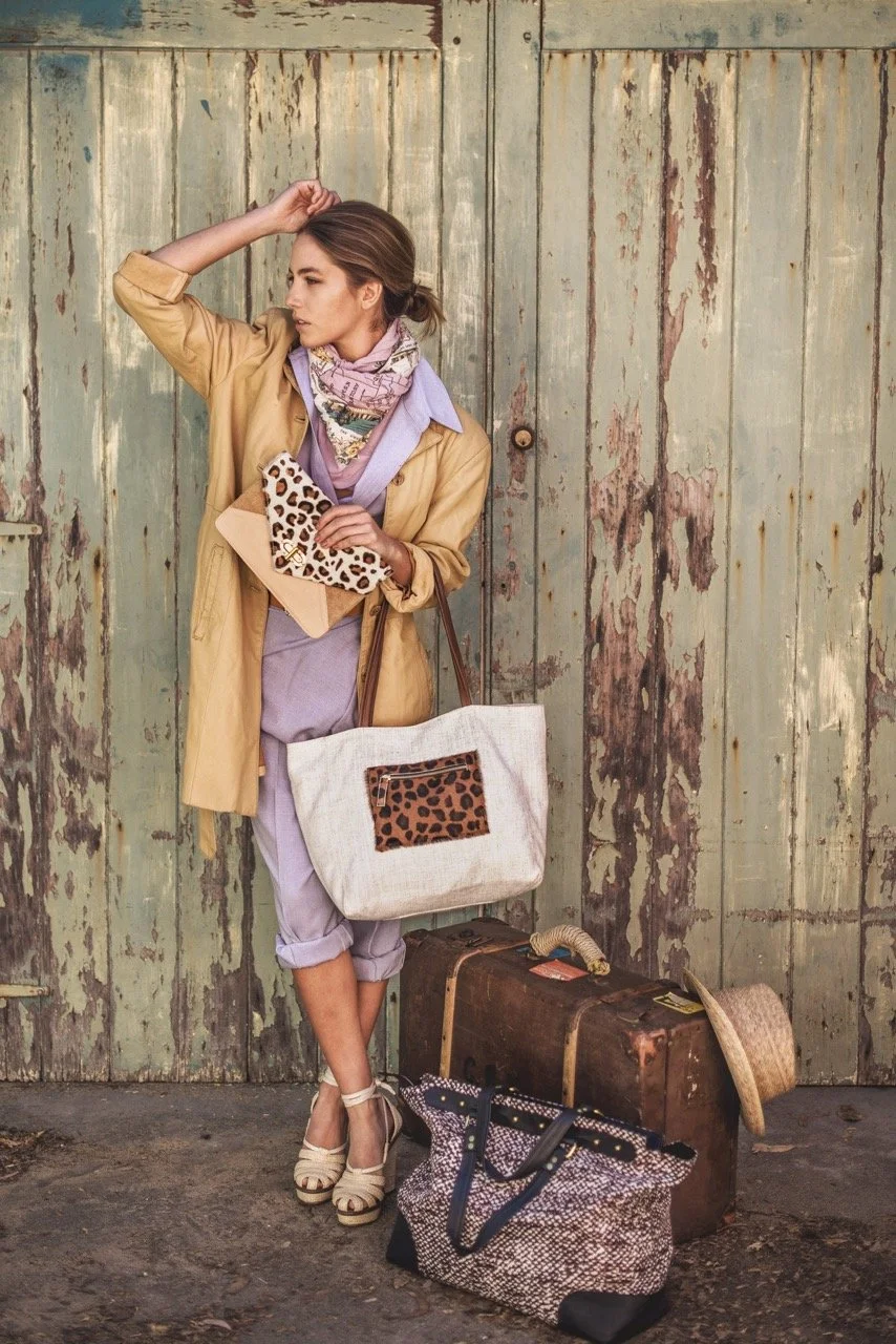 A woman standing in front of a weathered, green wooden door with peeling paint, dressed in a yellow coat, lavender jumpsuit, and beige high-heeled sandals. She is holding a leopard print clutch and has a large beige tote with a leopard patch, along w