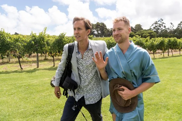 Two men walking in a vineyard, one in casual attire and the other in medical scrubs, engaged in conversation.